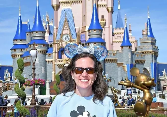 Woman smiling in front of Cinderella Castle at Walt Disney World, wearing Mickey Mouse ears and a graphic sweatshirt.